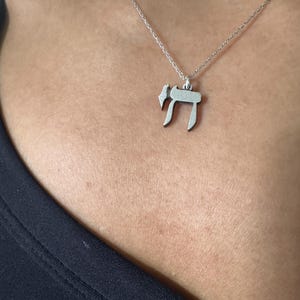 Puede incluir: Un collar de cadena de plata con un colgante que presenta la letra hebrea Jai, que simboliza la vida.