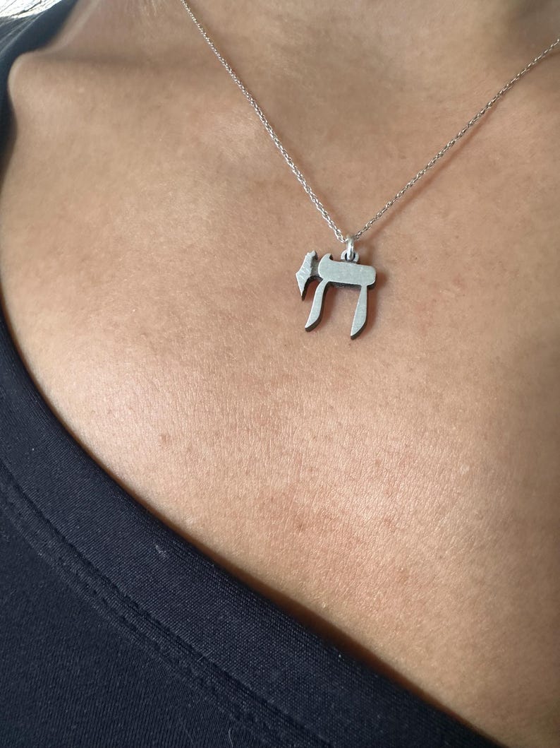 Puede incluir: Un collar de cadena de plata con un colgante que presenta la letra hebrea Jai, que simboliza la vida.