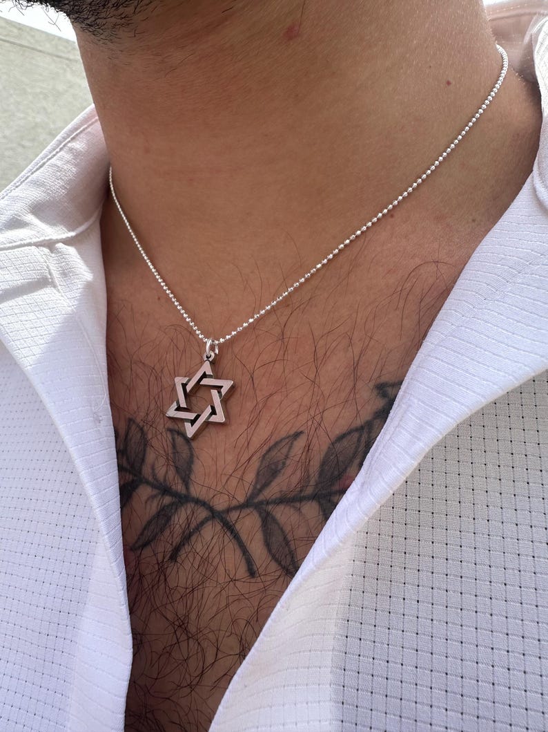 May include: A silver chain necklace with a silver Star of David pendant. The necklace is worn by a person with a tattoo of a branch on their chest.