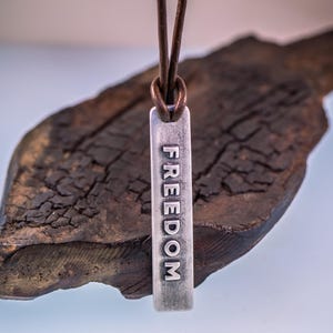 May include: A silver pendant necklace with the word "FREEDOM" engraved vertically. The pendant hangs from a brown leather cord. The background features a piece of dark, textured wood.