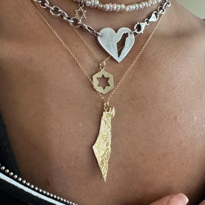 May include: A layered necklace set featuring a white pearl choker with a small Star of David charm, a silver chain with a heart pendant containing a map cutout, a gold chain with a hexagonal Star of David, and a gold textured map pendant.