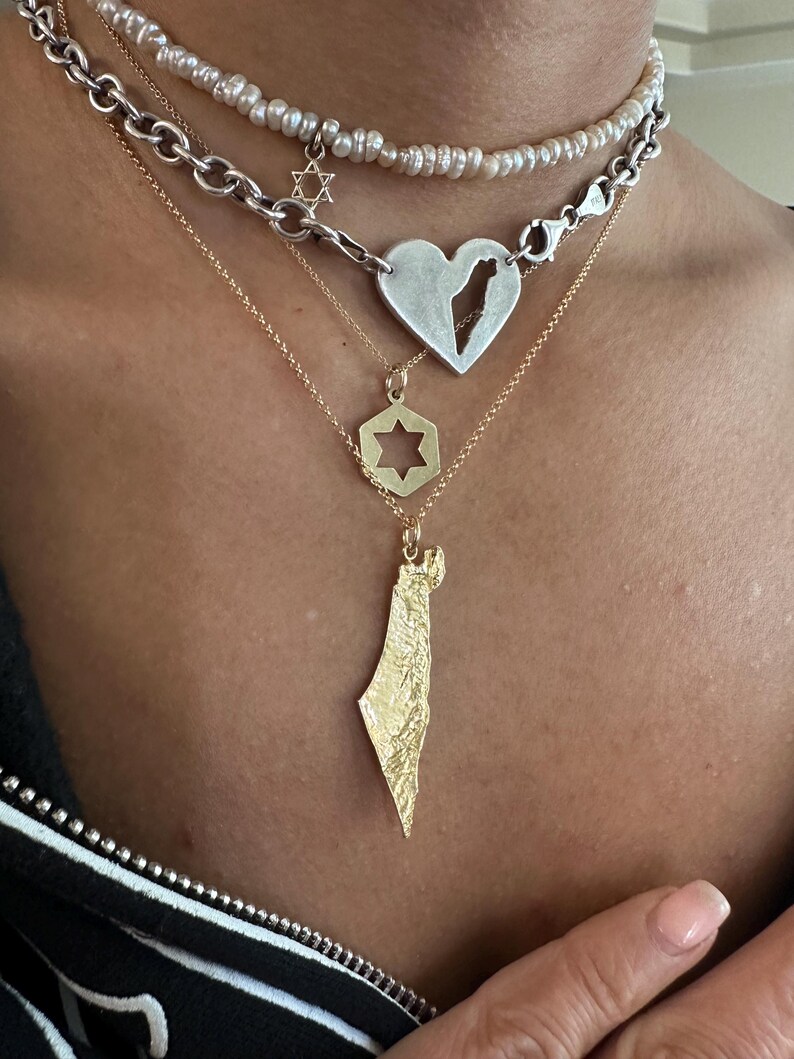 May include: A layered necklace set featuring a white pearl choker with a small Star of David charm, a silver chain with a heart pendant containing a map cutout, a gold chain with a hexagonal Star of David, and a gold textured map pendant.
