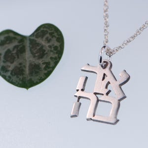 May include: A silver necklace with a pendant featuring the Japanese characters for "love" in a stylised font. The necklace is on a white surface next to a green heart-shaped leaf.