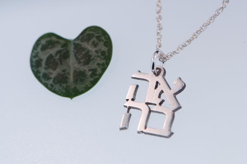 May include: A silver necklace with a pendant featuring the Japanese characters for "love" in a stylised font. The necklace is on a white surface next to a green heart-shaped leaf.
