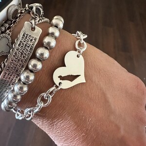 May include: A silver-toned bracelet featuring a heart-shaped charm with a cutout of a geographic landmass, connected to a thick chain link. Adjacent is a bracelet with large, round, polished silver-toned beads and a rectangular engraved metal tag.