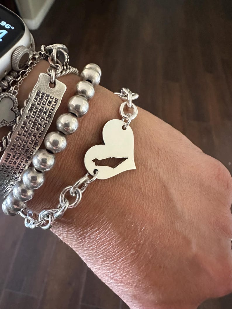 May include: A silver-toned bracelet featuring a heart-shaped charm with a cutout of a geographic landmass, connected to a thick chain link. Adjacent is a bracelet with large, round, polished silver-toned beads and a rectangular engraved metal tag.