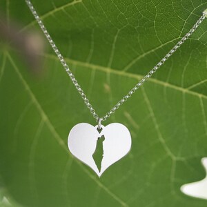 Op de afbeelding: Een zilveren ketting met een hartvormige hanger. De hanger heeft een uitgesneden ontwerp van de vorm van een land. De ketting wordt gepresenteerd tegen een groene bladeren achtergrond.