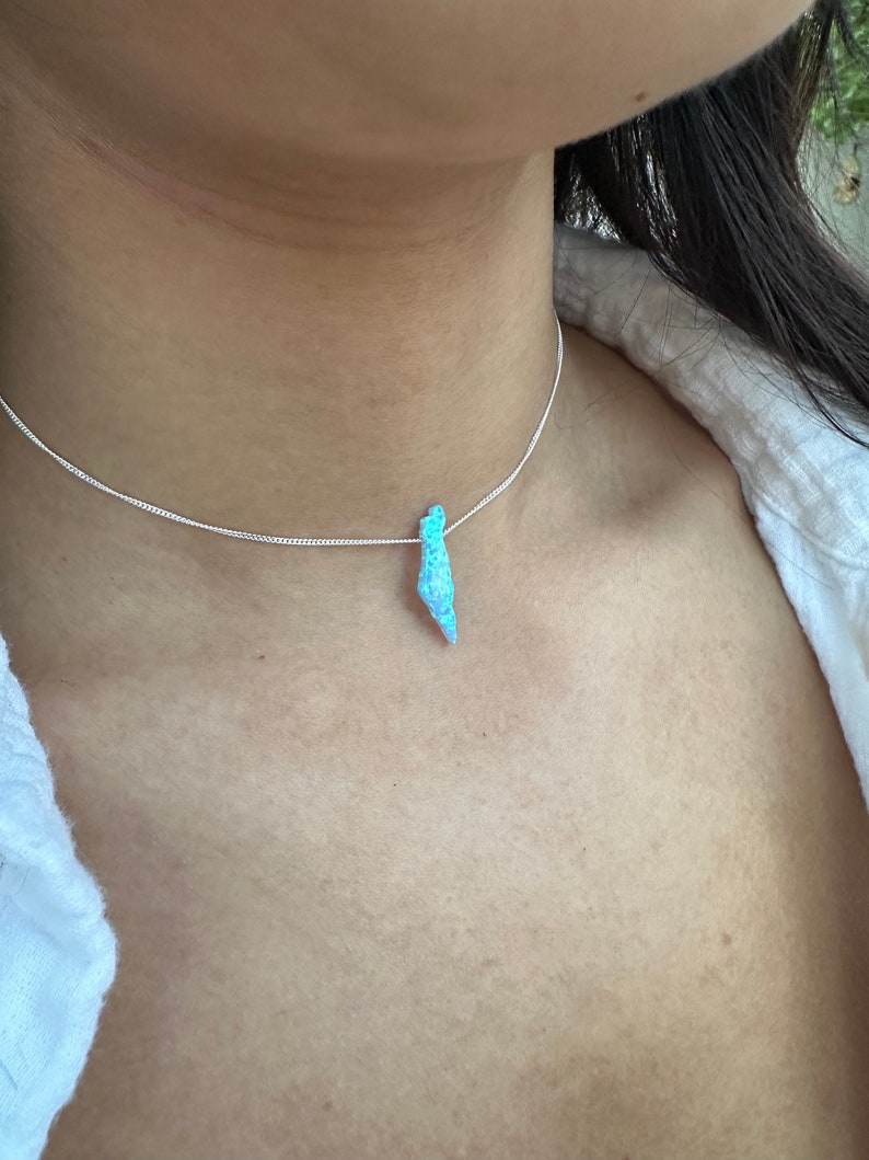 Op de afbeelding: Een zilveren ketting met een lichtblauwe opaal hanger. De opaal is druppelvormig en heeft een glinsterende, iriserende afwerking.