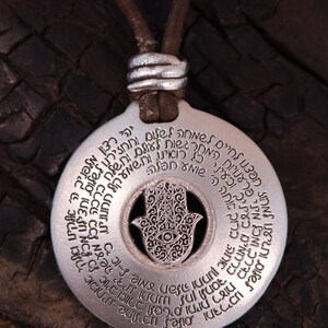 May include: A silver-colored pendant necklace with a brown leather cord. The pendant features a circular design with engraved text and a central Hamsa hand symbol with an eye. The necklace is a spiritual or religious item.