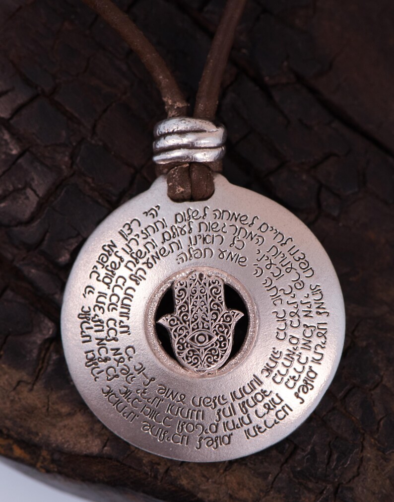 May include: A silver-colored pendant necklace with a brown leather cord. The pendant features a circular design with engraved text and a central Hamsa hand symbol with an eye. The necklace is a spiritual or religious item.