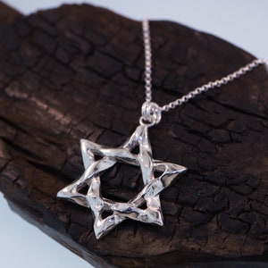 Puede incluir: Un collar con un colgante de plata en forma de Estrella de David con un dise&ntilde;o trenzado. El colgante est&aacute; colgado sobre una superficie de madera oscura.