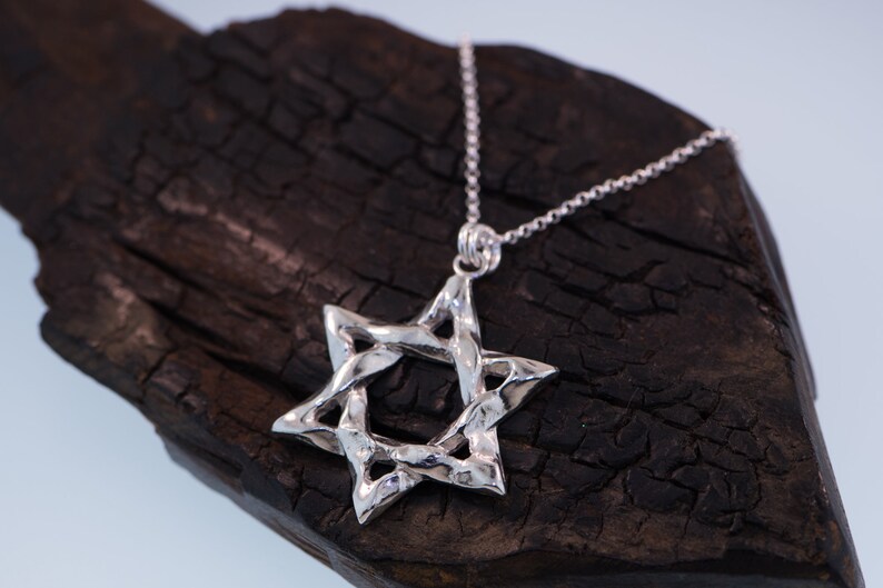 Puede incluir: Un collar con un colgante de plata en forma de Estrella de David con un dise&ntilde;o trenzado. El colgante est&aacute; colgado sobre una superficie de madera oscura.