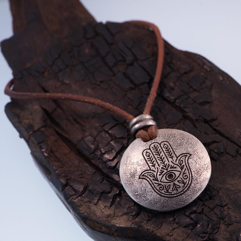 May include: A silver-colored pendant necklace featuring a Hamsa hand design with an eye in the center. The pendant hangs from a brown leather cord. The necklace is displayed on a dark, textured wooden surface.