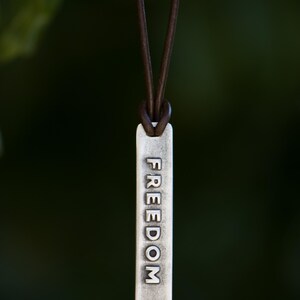 May include: A silver-colored rectangular pendant with the word "FREEDOM" engraved on it, suspended from a brown leather cord. The pendant hangs against a blurred green background.