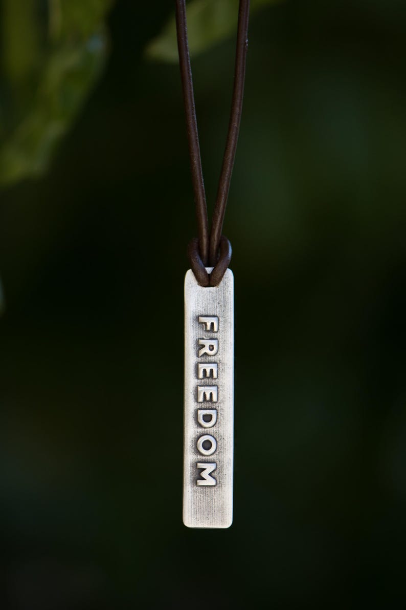 May include: A silver-colored rectangular pendant with the word "FREEDOM" engraved on it, suspended from a brown leather cord. The pendant hangs against a blurred green background.