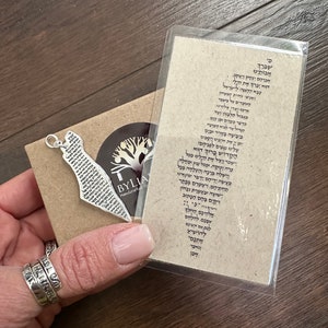 May include: A silver-colored pendant in the shape of a map, with Hebrew text, rests on a small brown card. The card is next to a clear plastic sleeve with more Hebrew text. The pendant is on a brown card with a logo that says "BYLIA Handcrafted".