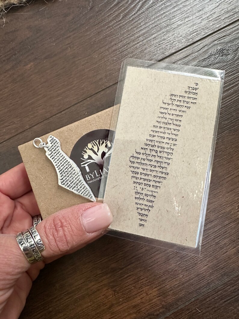 May include: A silver-colored pendant in the shape of a map, with Hebrew text, rests on a small brown card. The card is next to a clear plastic sleeve with more Hebrew text. The pendant is on a brown card with a logo that says "BYLIA Handcrafted".