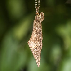 May include: A gold-toned metal pendant shaped like the map of Israel, featuring a textured, topographical surface detail. The pendant hangs from a delicate matching gold-toned metal chain, suspended vertically against a blurred green background.