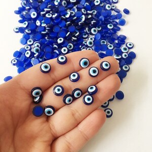 May include: A close-up of a hand holding a collection of small blue and white evil eye charms. The charms are round and have a white center with a blue eye.