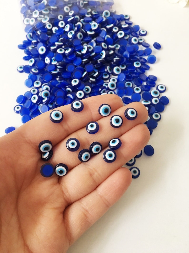 May include: A close-up of a hand holding a collection of small blue and white evil eye charms. The charms are round and have a white center with a blue eye.