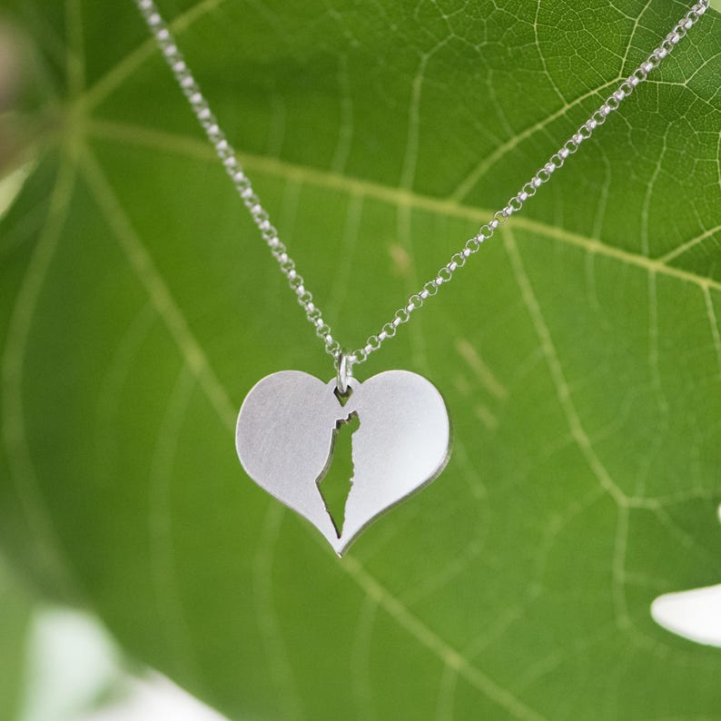 Op de afbeelding: Zilveren ketting met een hartvormige hanger. De hanger heeft een uitgesneden ontwerp van de kaart van een land. De ketting hangt tegen een groene bladeren achtergrond.