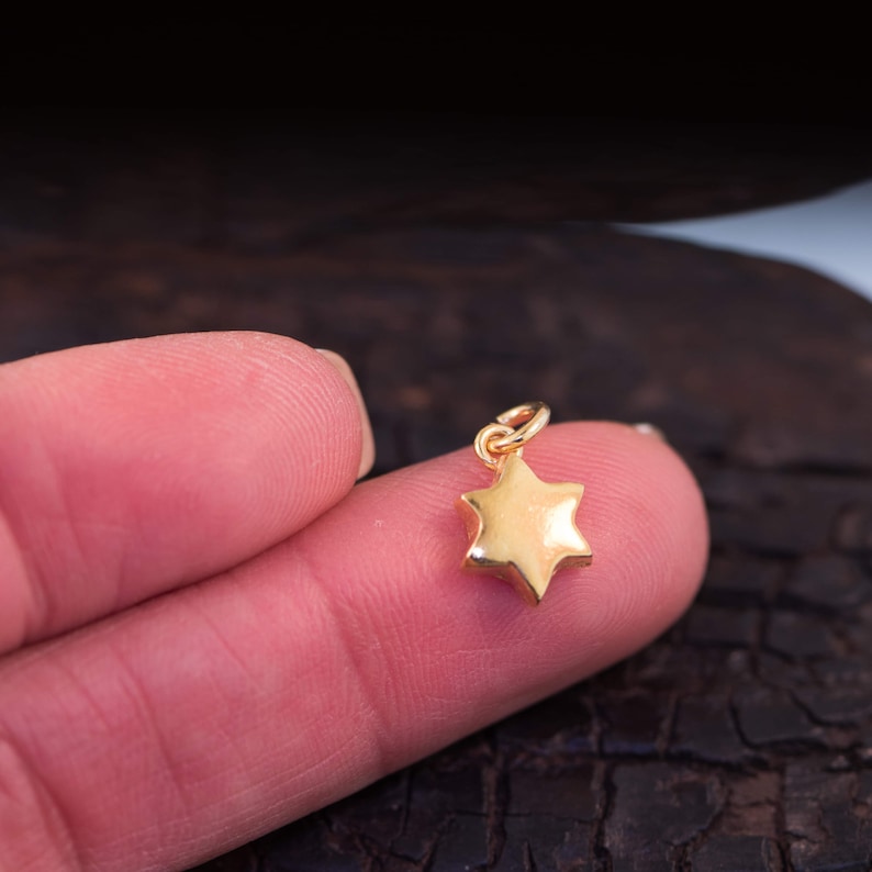 May include: A small, gold-coloured star charm with six points. The charm has a loop at the top for attaching to a necklace or bracelet. The star is held between two fingers.