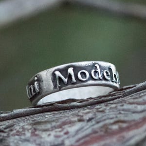 Puede incluir: Un anillo de plata con las palabras "ini Model" grabadas alrededor de la banda. El anillo se encuentra sobre un fondo natural borroso, resaltando la inscripci&oacute;n y el brillo met&aacute;lico del anillo. El anillo es una joya.