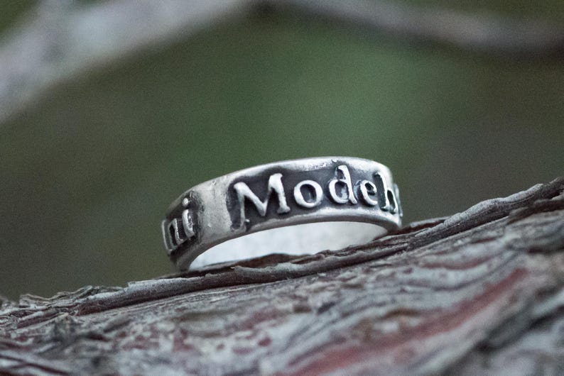 Puede incluir: Un anillo de plata con las palabras "ini Model" grabadas alrededor de la banda. El anillo se encuentra sobre un fondo natural borroso, resaltando la inscripci&oacute;n y el brillo met&aacute;lico del anillo. El anillo es una joya.