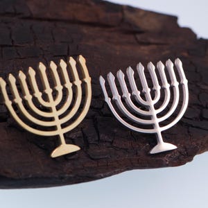 K&ouml;nnte beinhalten: Zwei Menora-Anh&auml;nger, einer goldfarben und einer silberfarben, liegen auf einer dunklen, strukturierten Holzoberfl&auml;che. Die Menoras haben ein identisches Design mit neun Kerzenhaltern. Der Hintergrund ist ein neutrales, helles Blau.