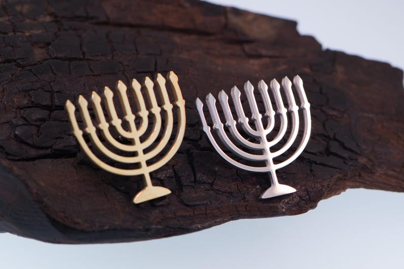 K&ouml;nnte beinhalten: Zwei Menora-Anh&auml;nger, einer goldfarben und einer silberfarben, liegen auf einer dunklen, strukturierten Holzoberfl&auml;che. Die Menoras haben ein identisches Design mit neun Kerzenhaltern. Der Hintergrund ist ein neutrales, helles Blau.