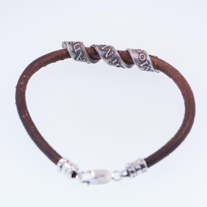 May include: A brown leather bracelet with three silver-coloured, spiralled bands. Each band has engraved text. The bracelet has a silver clasp.