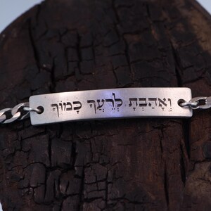 Op de afbeelding: Een zilverkleurige armband met een rechthoekige plaat gegraveerd met Hebreeuwse tekst. De armband heeft een schakelketting ontwerp. De plaat is met kleine lussen aan de ketting bevestigd. De achtergrond is een donker, getextureerd houten oppervlak.