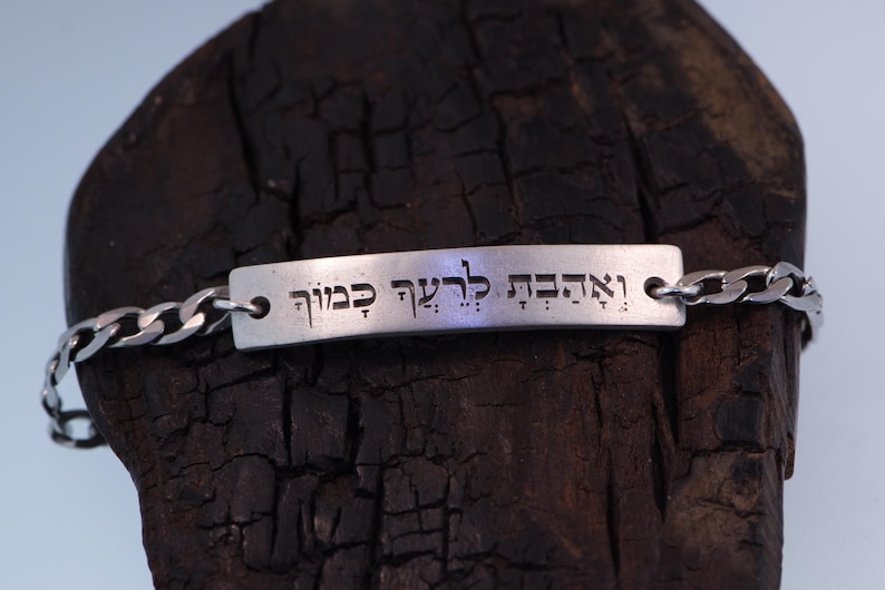 Op de afbeelding: Een zilverkleurige armband met een rechthoekige plaat gegraveerd met Hebreeuwse tekst. De armband heeft een schakelketting ontwerp. De plaat is met kleine lussen aan de ketting bevestigd. De achtergrond is een donker, getextureerd houten oppervlak.