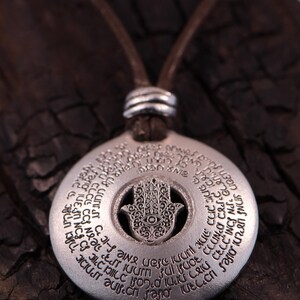 May include: Silver-colored pendant necklace featuring a Hamsa hand design with an eye in the center. The pendant has engraved text around the edge and hangs from a brown leather cord. The necklace is displayed on a dark, textured surface.