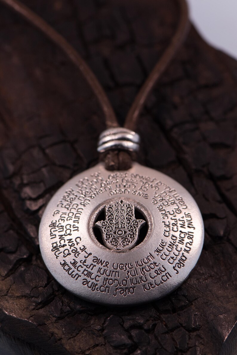 May include: Silver-colored pendant necklace featuring a Hamsa hand design with an eye in the center. The pendant has engraved text around the edge and hangs from a brown leather cord. The necklace is displayed on a dark, textured surface.