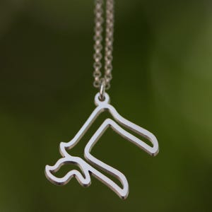 Peut inclure: Un collier pendentif argent&eacute; avec un design abstrait unique. Le pendentif pr&eacute;sente une forme stylis&eacute;e avec des &eacute;l&eacute;ments courbes et angulaires, suspendu &agrave; une cha&icirc;ne d&eacute;licate. L'arri&egrave;re-plan est vert flou.