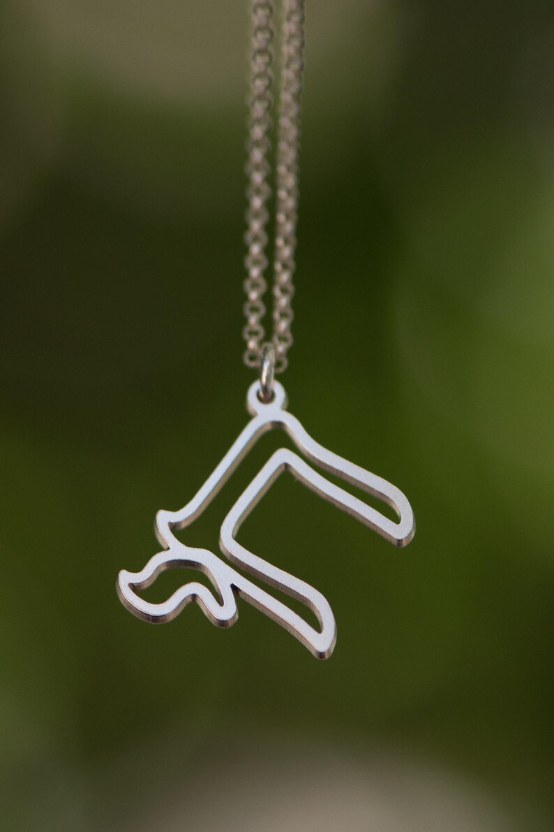 Peut inclure: Un collier pendentif argent&eacute; avec un design abstrait unique. Le pendentif pr&eacute;sente une forme stylis&eacute;e avec des &eacute;l&eacute;ments courbes et angulaires, suspendu &agrave; une cha&icirc;ne d&eacute;licate. L'arri&egrave;re-plan est vert flou.