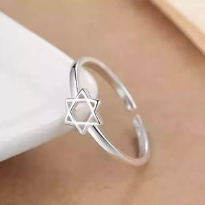 Puede incluir: Un anillo abierto de tono plateado que presenta un emblema de la Estrella de David de seis puntas. El anillo tiene un acabado met&aacute;lico pulido y un dise&ntilde;o de banda delgada y simple que es ajustable, permitiendo un ajuste flexible en varios tama&ntilde;os.
