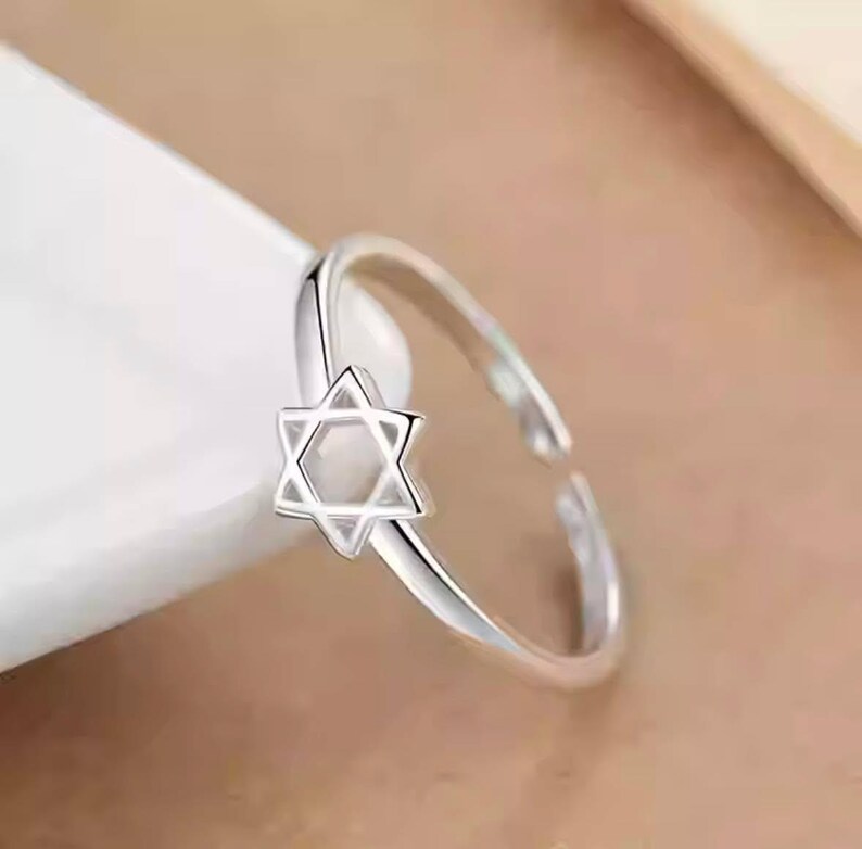 Puede incluir: Un anillo abierto de tono plateado que presenta un emblema de la Estrella de David de seis puntas. El anillo tiene un acabado met&aacute;lico pulido y un dise&ntilde;o de banda delgada y simple que es ajustable, permitiendo un ajuste flexible en varios tama&ntilde;os.