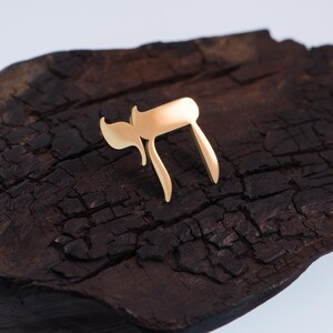 K&ouml;nnte beinhalten: Ein goldfarbenes Chai-Symbol, das im Hebr&auml;ischen "Leben" bedeutet, liegt auf einer dunklen, strukturierten Holzoberfl&auml;che. Das Design des Symbols zeigt zwei hebr&auml;ische Buchstaben, die eine stilisierte Darstellung bilden. Der Hintergrund ist dunkles, raues Holz.