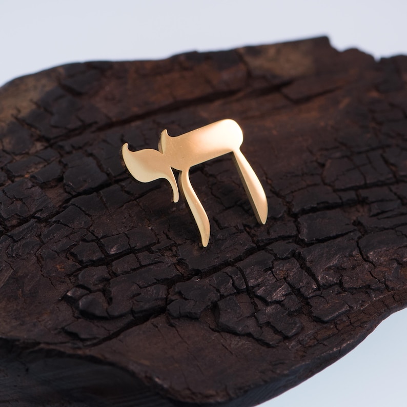 K&ouml;nnte beinhalten: Ein goldfarbenes Chai-Symbol, das im Hebr&auml;ischen "Leben" bedeutet, liegt auf einer dunklen, strukturierten Holzoberfl&auml;che. Das Design des Symbols zeigt zwei hebr&auml;ische Buchstaben, die eine stilisierte Darstellung bilden. Der Hintergrund ist dunkles, raues Holz.