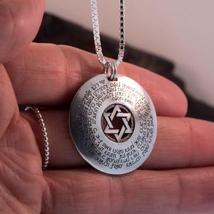 Puede incluir: Un collar con colgante de plata con un dije circular grabado con texto y una Estrella de David en el centro. El colgante cuelga de una cadena fina. El collar se sostiene en una mano.