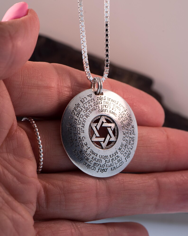 Puede incluir: Un collar con colgante de plata con un dije circular grabado con texto y una Estrella de David en el centro. El colgante cuelga de una cadena fina. El collar se sostiene en una mano.
