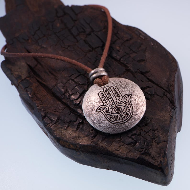May include: A silver-colored pendant necklace featuring a Hamsa hand design with an eye in the center. The pendant hangs from a brown leather cord. The necklace is displayed on a dark, textured wooden surface.
