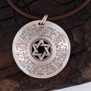 Puede incluir: Collar con colgante de plata con un dise&ntilde;o de Estrella de David en el centro, rodeado de texto hebreo. El colgante cuelga de un cord&oacute;n de cuero marr&oacute;n. El fondo es una superficie oscura y texturizada.