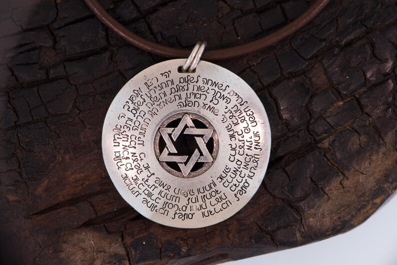 Puede incluir: Collar con colgante de plata con un dise&ntilde;o de Estrella de David en el centro, rodeado de texto hebreo. El colgante cuelga de un cord&oacute;n de cuero marr&oacute;n. El fondo es una superficie oscura y texturizada.