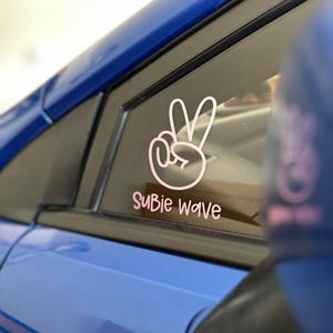 以下が含まれることがあります： ピンク色のピースサインと「Subie Wave」の文字が入ったステッカーが貼られた青い車の窓。ステッカーは車の窓にあり、背景にはサイドミラーが見えます。