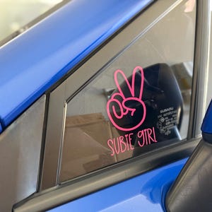 May include: A pink peace sign sticker on a blue car window with the text "SUBIE GIRL" below it.