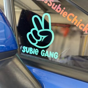 Puede incluir: Una pegatina de color azul turquesa en forma de signo de paz con el texto "Subie Gang" en la ventana de un coche. La pegatina está en un coche azul.