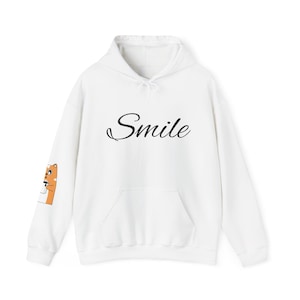 May include: White hoodie with a front pocket and drawstring. The word "Smile" is written in cursive across the chest. A cartoon graphic is on the left sleeve. This casual sweatshirt is perfect for everyday wear.